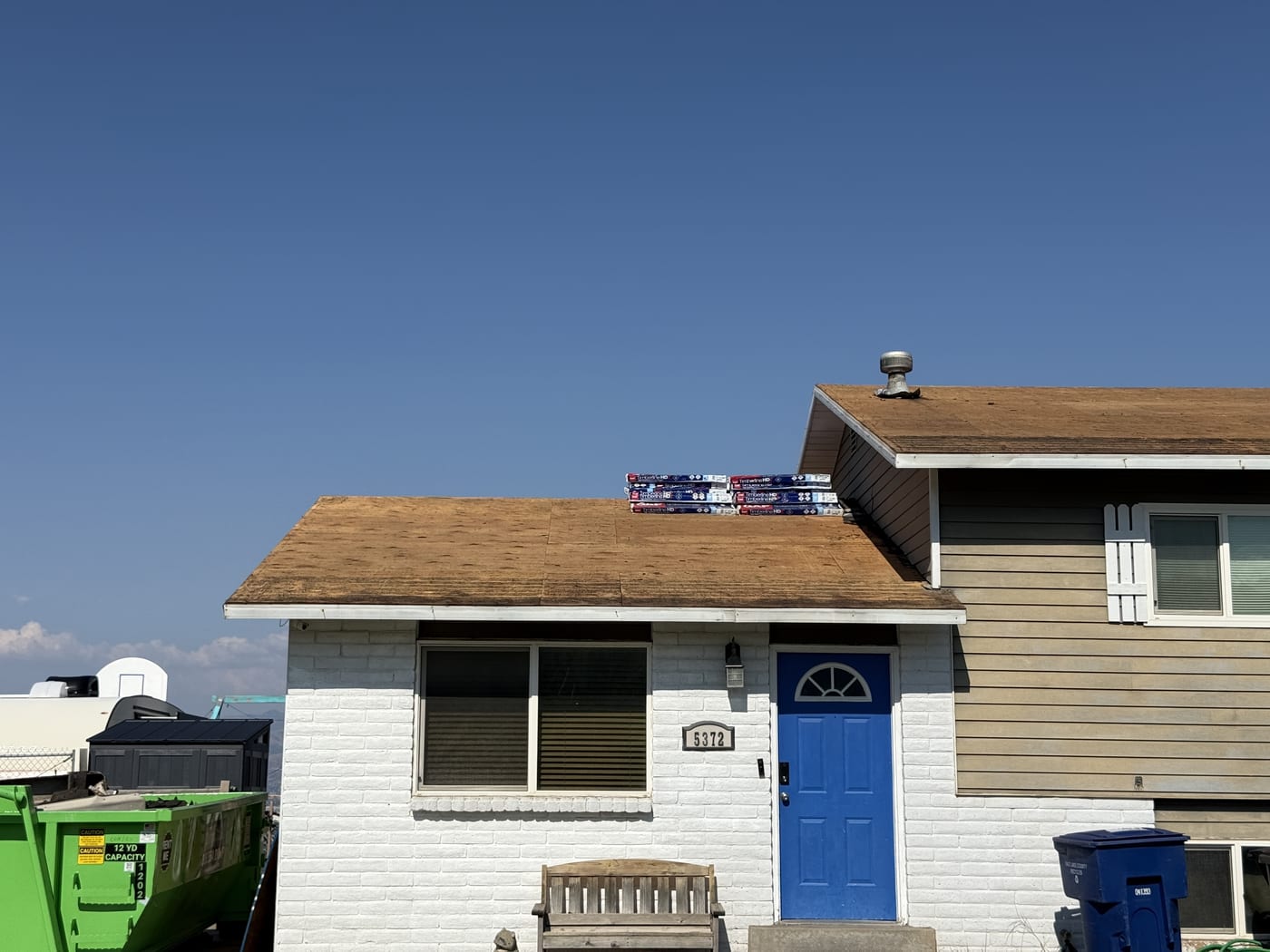 View of house exterior with blue front door (#5372), bare OSB roof deck with shingle bundles, dumpster visible at right