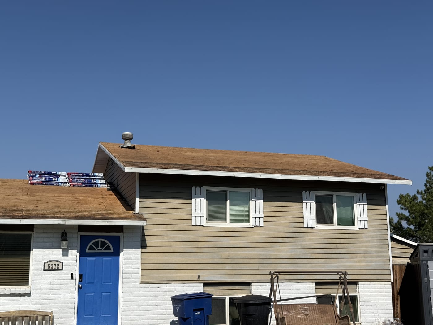 View of house exterior (blue door, brick siding), bare OSB roof deck with shingle bundles, in-progress re-roof