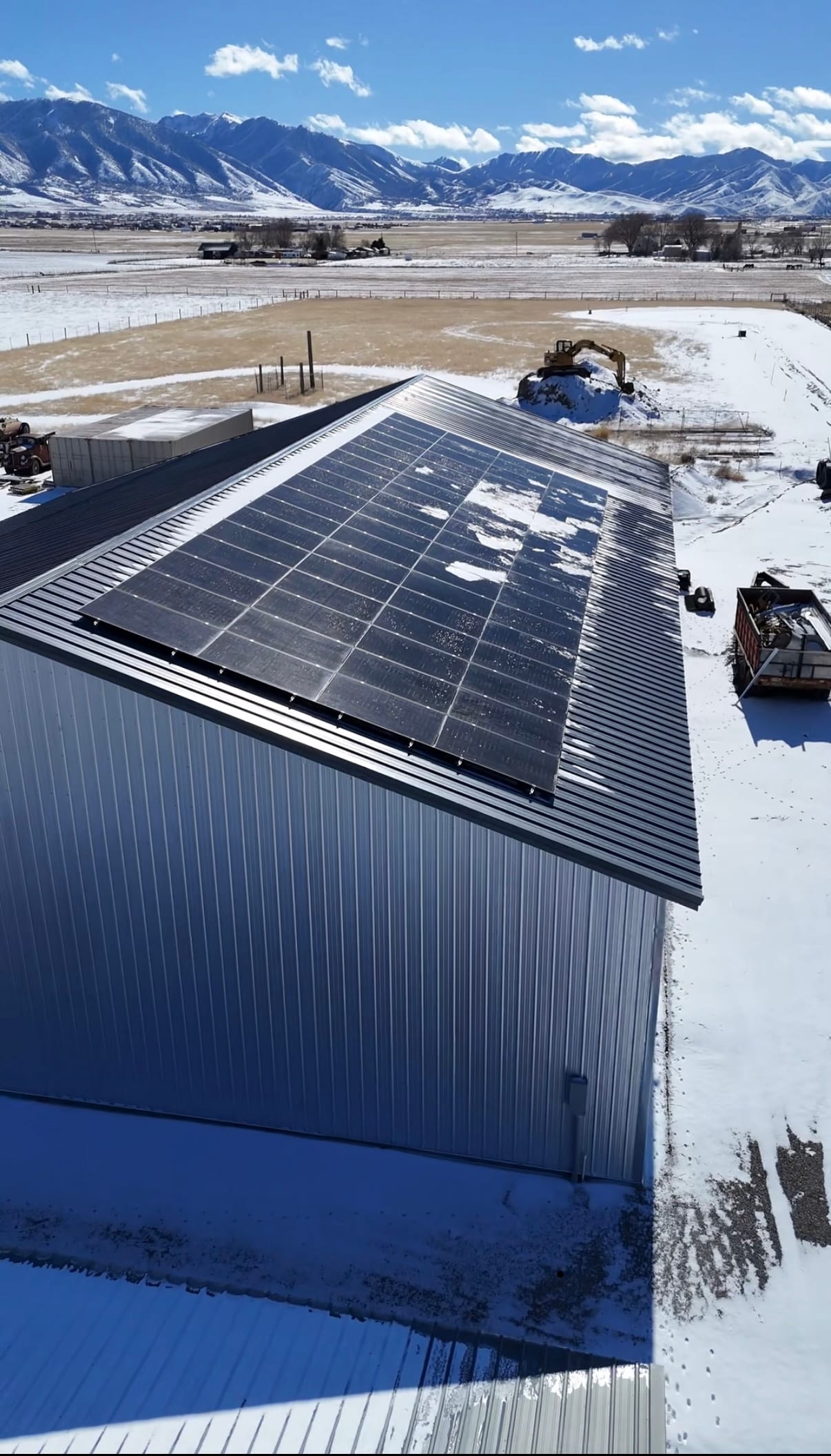 [Tier 1 Trina panels] Drone aerial of large metal barn/outbuilding with solar panels on roof, mountain backdrop, snow on ground, and construction equipment visible nearby