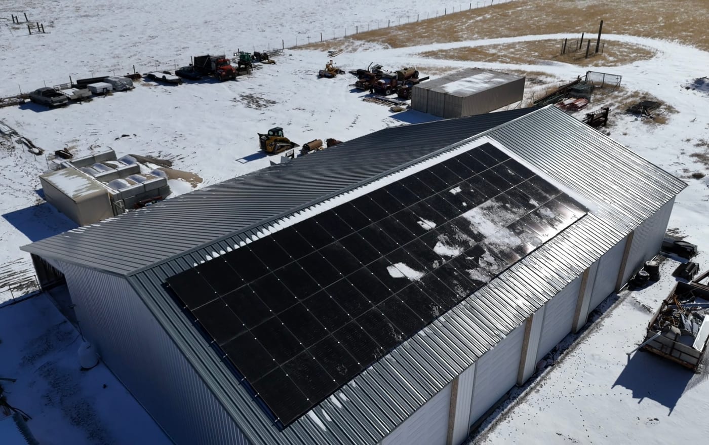 [Tier 1 Trina panels] Overhead drone shot of large metal agricultural building with rooftop solar array, snow-covered property, and heavy equipment in yard