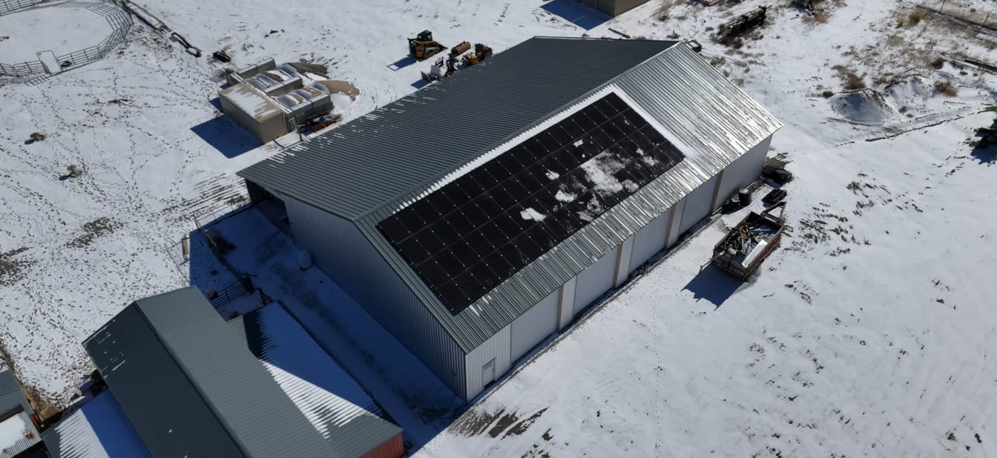 [Tier 1 Trina panels] Direct overhead drone shot of large metal agricultural building with rooftop solar panels and snow-covered property