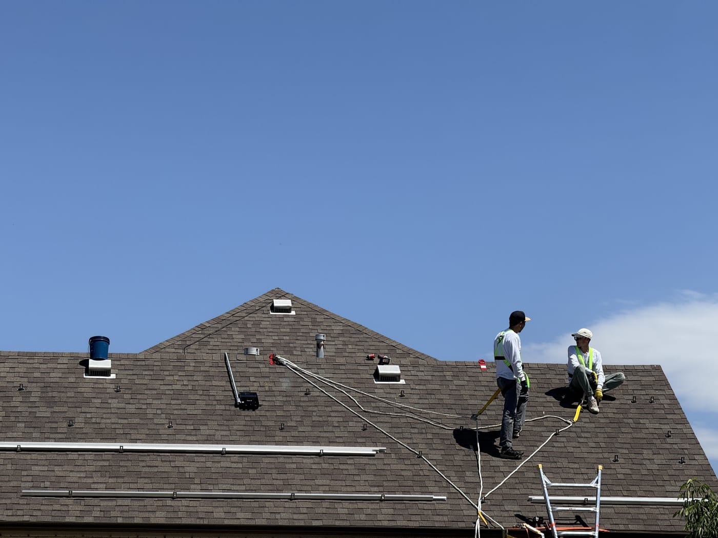 Two crew members with safety harnesses installing racking rails on shingle roof with ladder and wiring visible