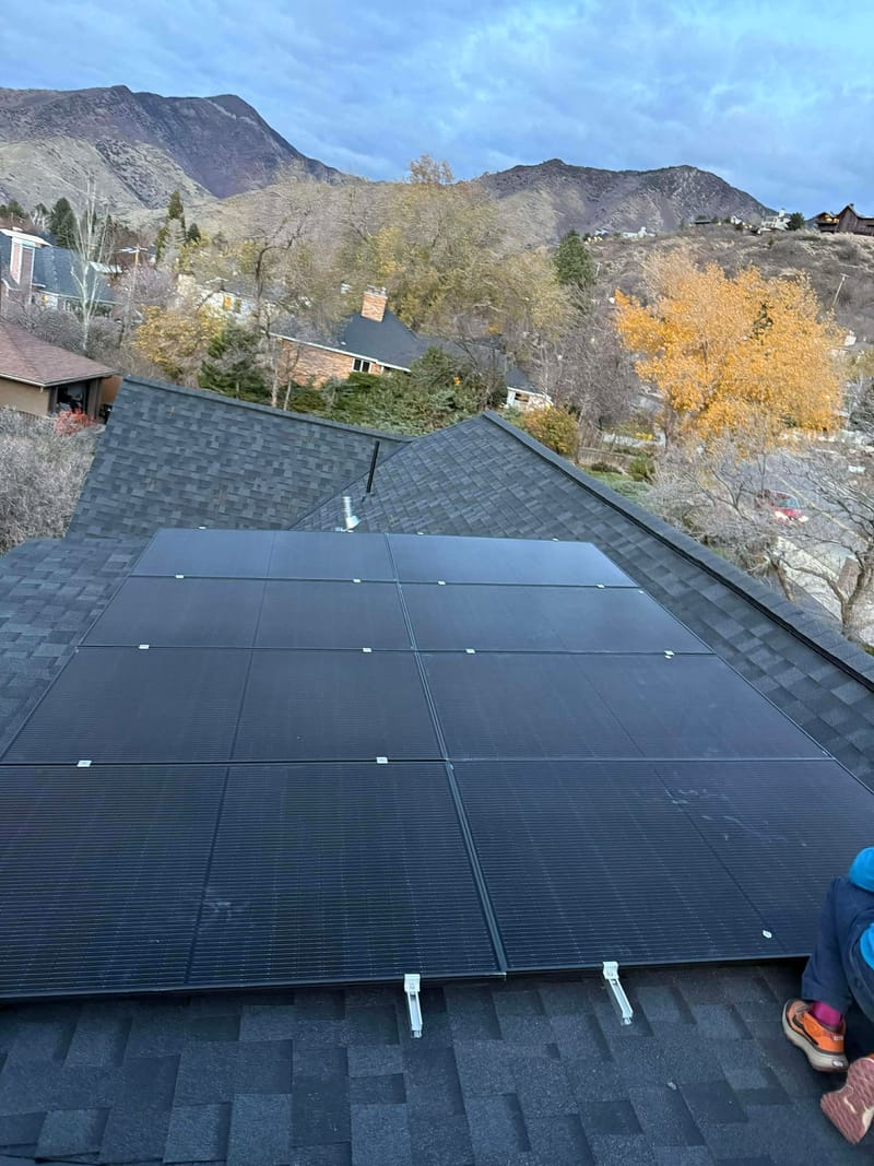 Black solar panels on gray shingle roof with fall foliage and Wasatch mountains behind, shot from roof level
