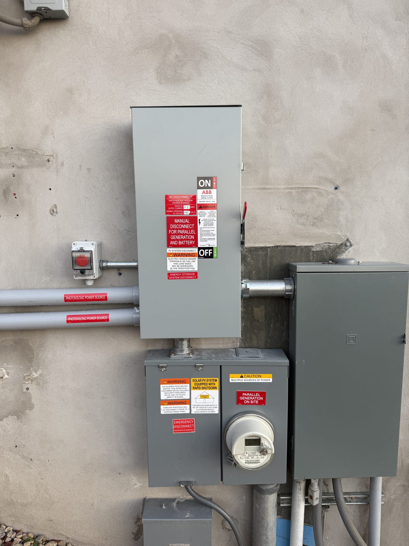 Wider view of exterior electrical equipment showing disconnect, meter, conduit runs, and warning labels on concrete wall