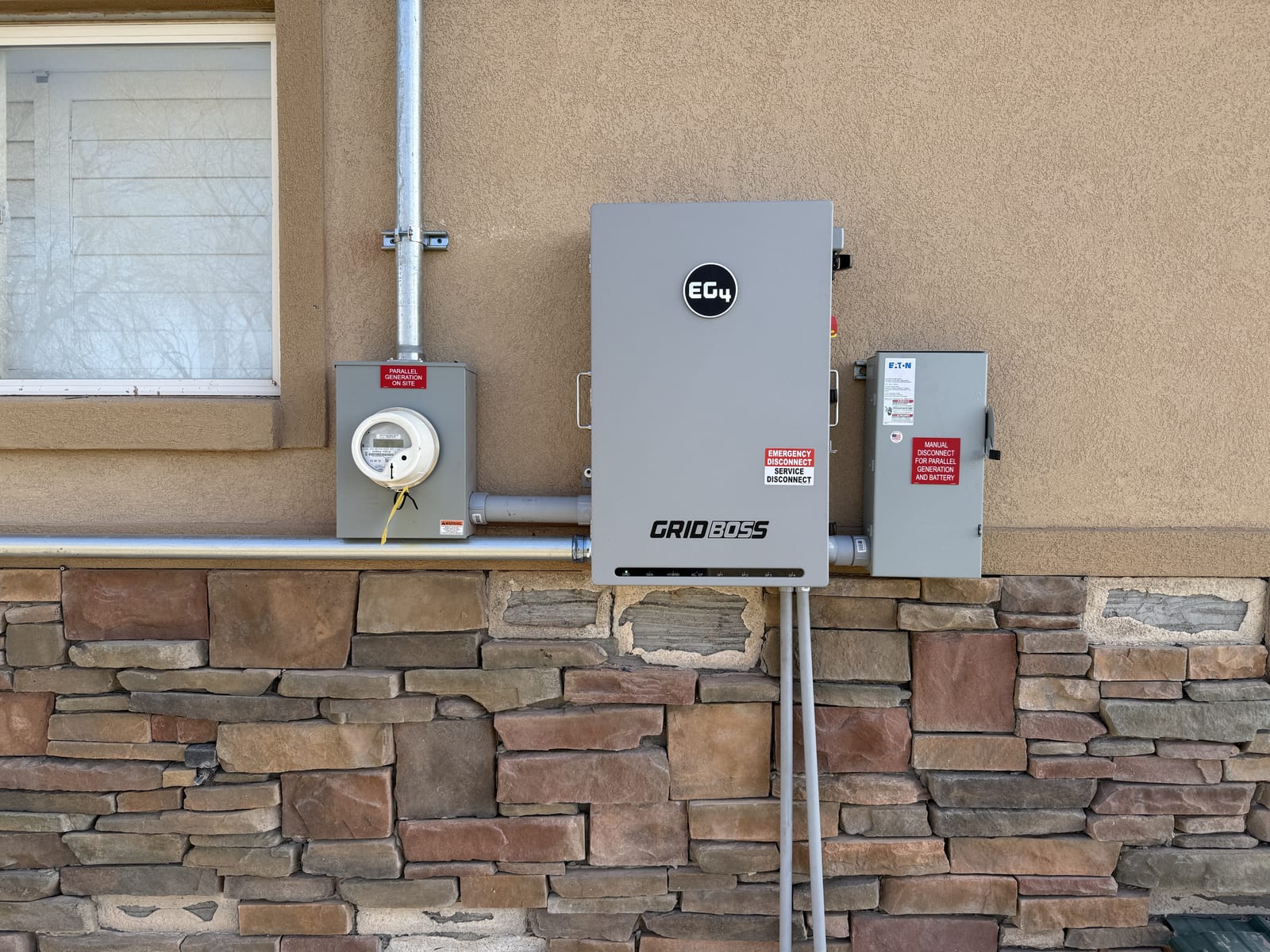 EG4 GridBOSS inverter mounted on exterior stone/stucco wall with meter socket and disconnect, conduit runs visible