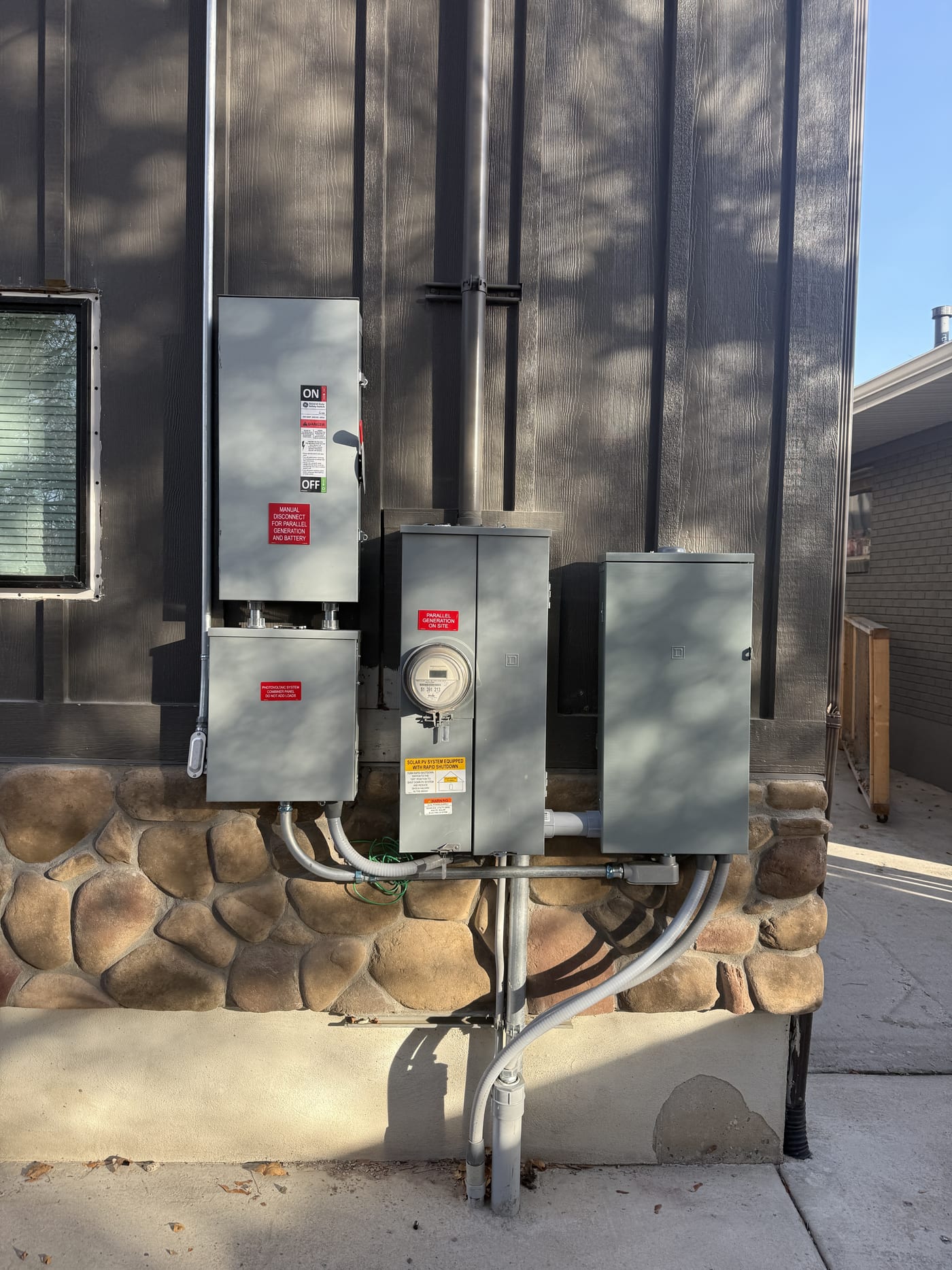 Wider view of exterior electrical equipment including meter, panels, disconnects, and conduit with basement window visible