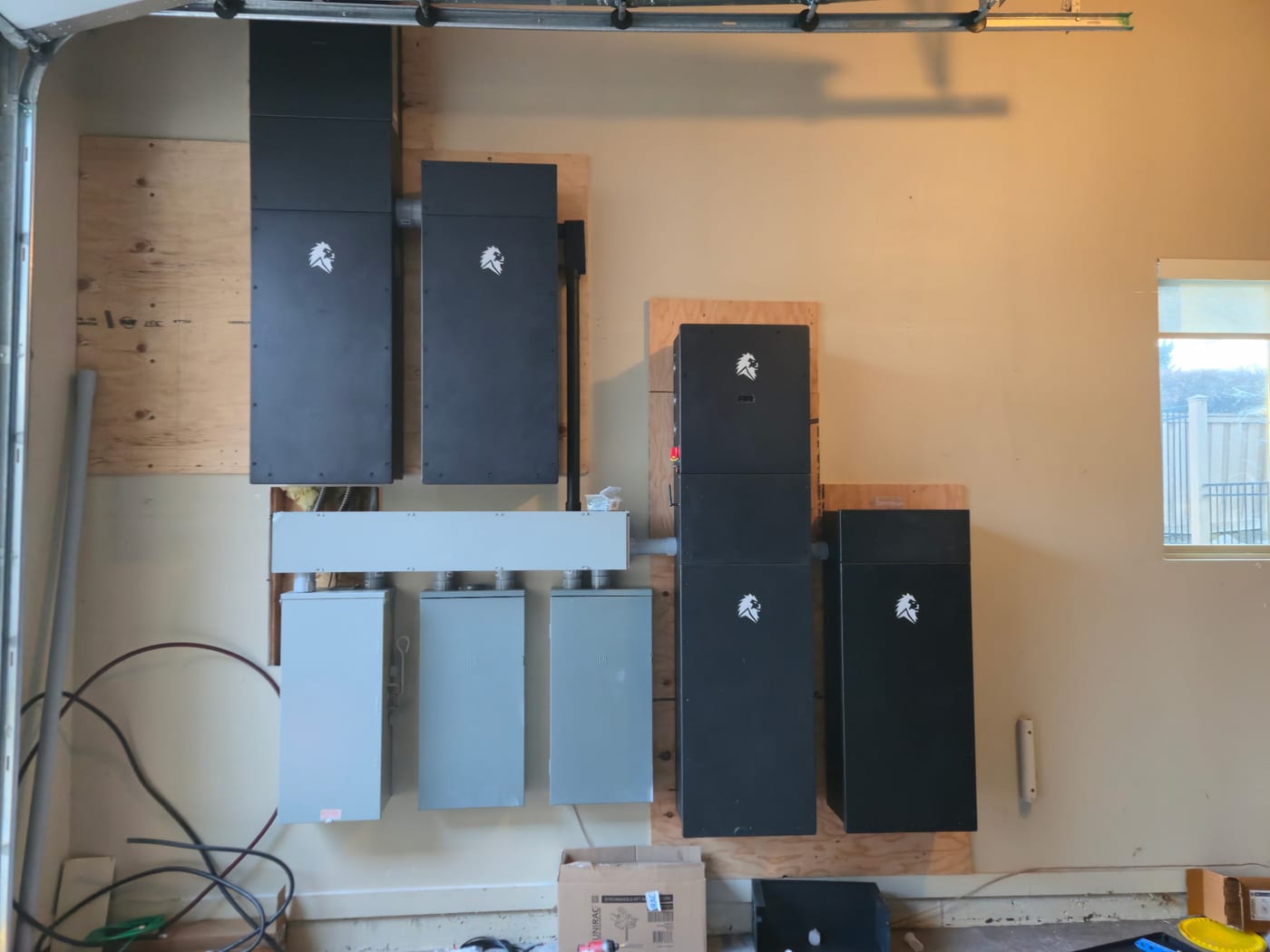 Lion Energy Sanctuary battery system fully mounted in garage — four large black battery cabinets with white lion logos arranged around three gray Lion Energy disconnect panels and white busbar/junction enclosure, plywood backboards (image rotated)