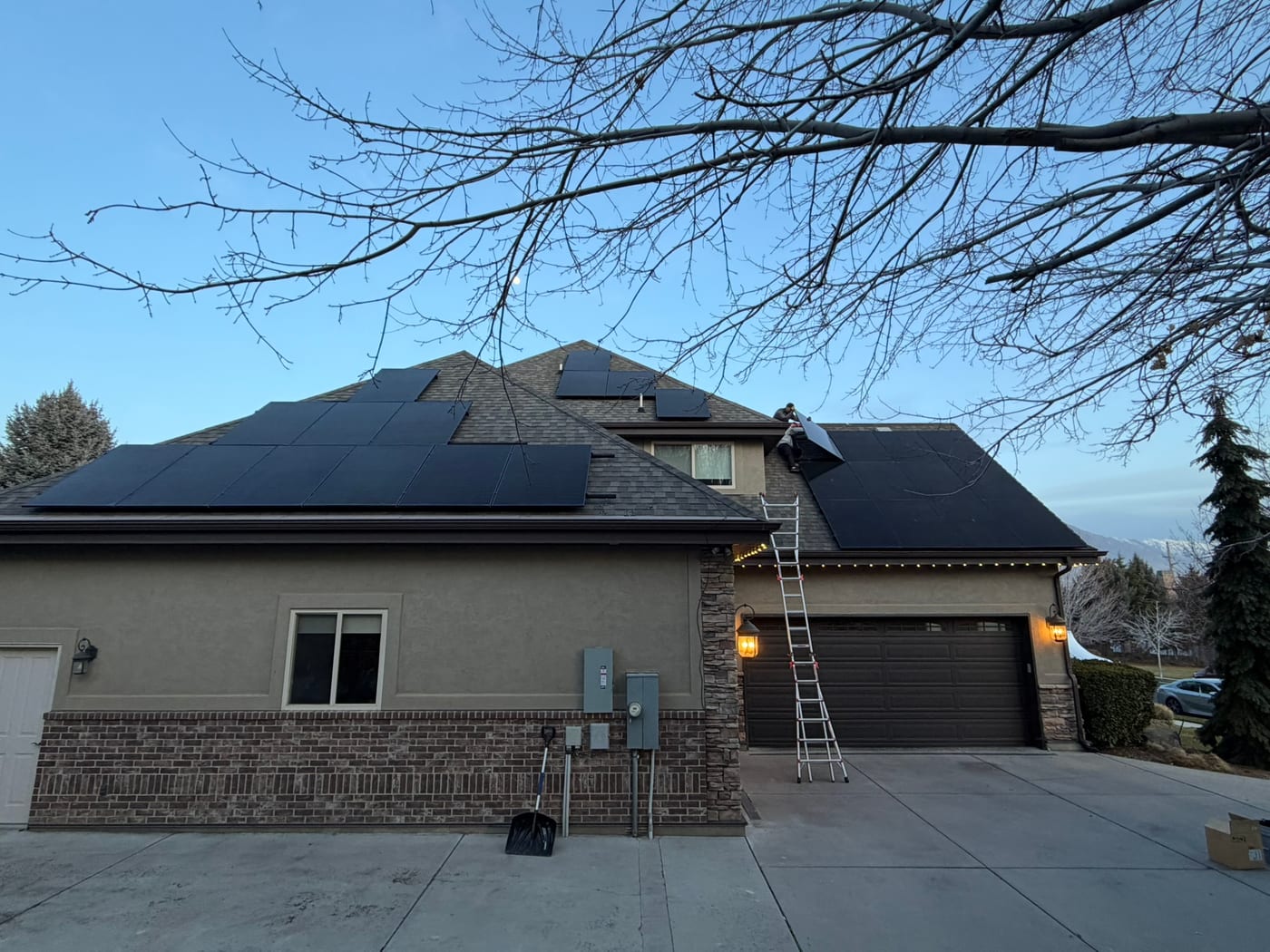 Front view of large dark stone and stucco home with solar panels on roof, ladder against house indicating active installation, driveway and garage visible